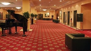 Un vestíbulo de hotel con un piano de cola, alfombra roja y un largo pasillo con puertas y una recepción.