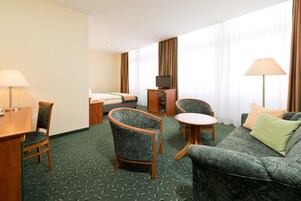 Habitación de hotel con cama, escritorio con lámpara, dos sillones, sofá y televisor sobre un mueble.