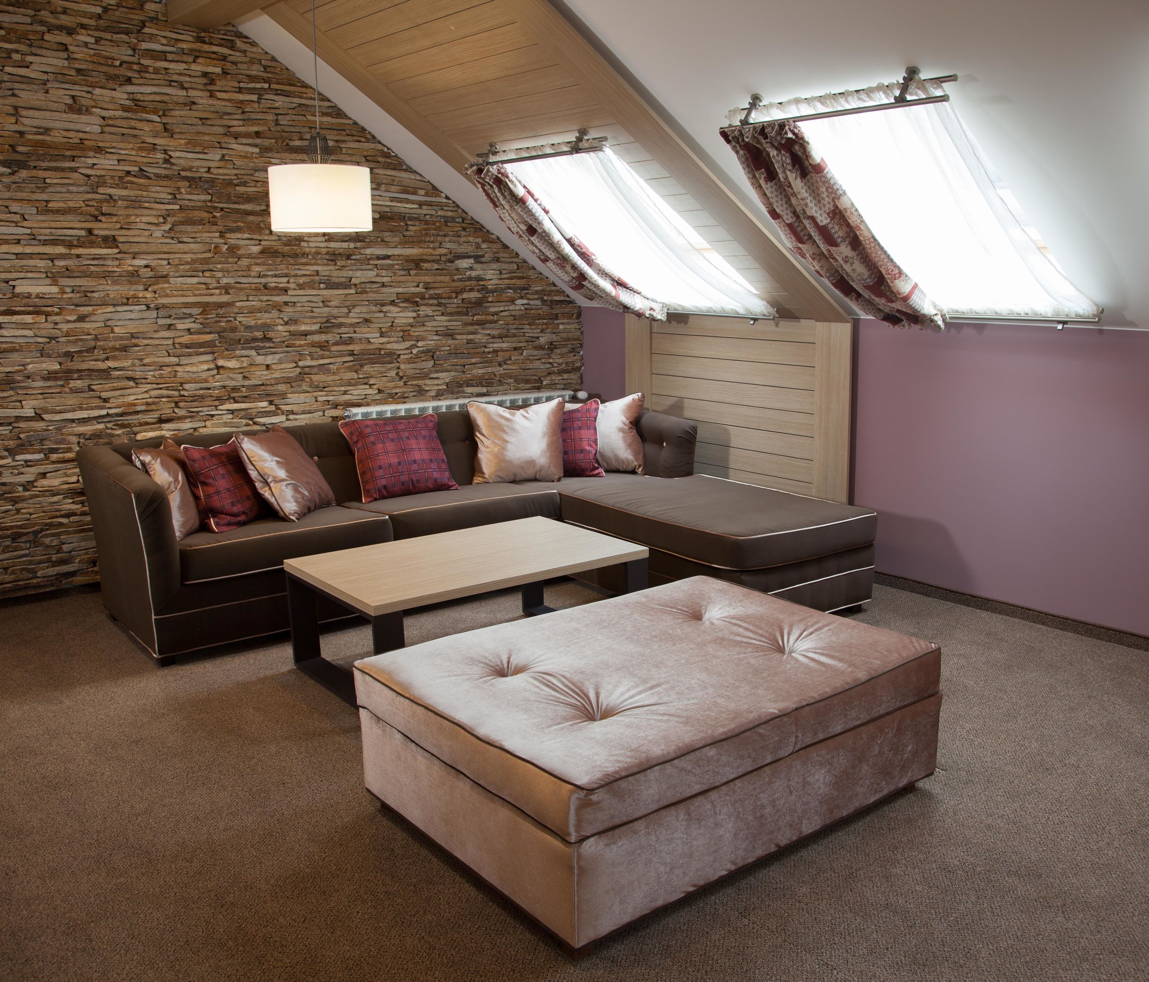 A loft-style room with a stone wall, a sectional sofa with patterned cushions, a small ottoman, and a wooden headboard.