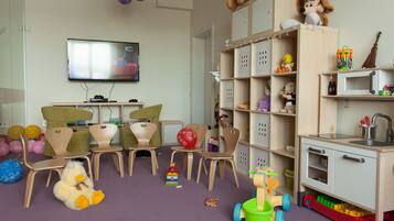 Una sala de juegos infantiles con televisor, sillas para jugar, un juego de cocina y varios juguetes.