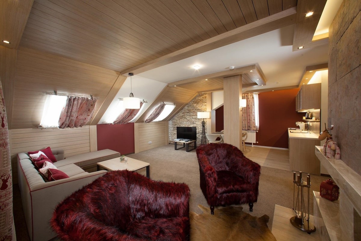 A cozy loft-style living space with a high ceiling, sloped roof, and skylights. It features a sofa, a red furry chair, a dining table, and a kitchen area with a sink and cabinets.