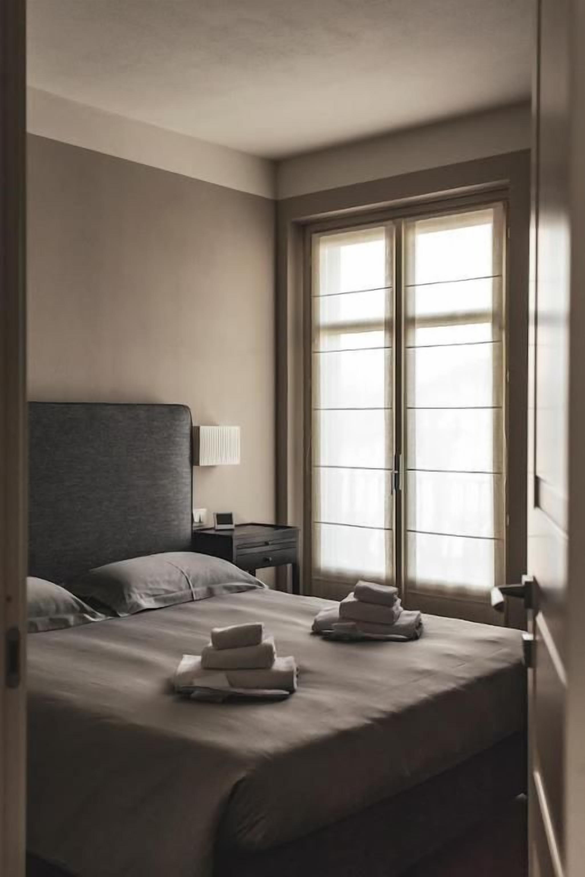 A hotel room with a bed, a nightstand, a lamp, and a window with blinds.
