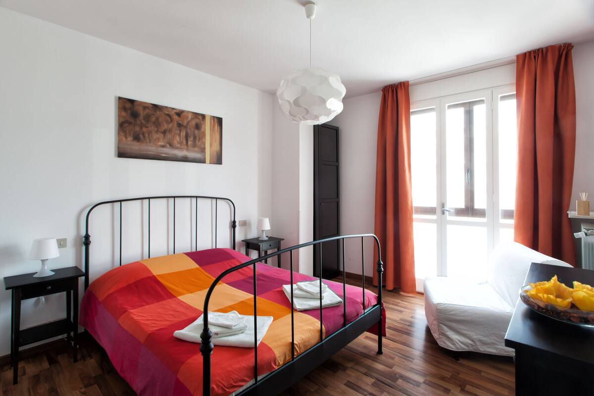 Un lit bien fait, agrémenté d’une couvre-lit aux couleurs vives, une table de chevet avec une lampe, un tableau accroché au mur et une fenêtre avec des rideaux.