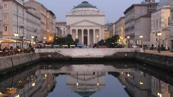 A historic building with a central dome and columns, reflecting in a calm water body, surrounded by urban architecture.