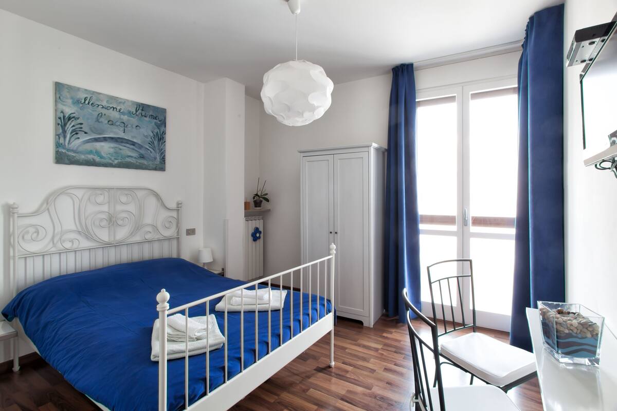 Une chambre avec un lit en métal blanc, une couvre-lit bleue, un radiateur blanc, une armoire blanche, une chaise blanche, une table blanche et un rideau bleu.