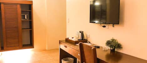 Una habitación pequeña con un escritorio de madera, una silla, un televisor, un cesto de basura y una planta.