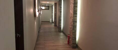 A long, narrow hallway with wooden flooring and walls on both sides. There are doors on the left and a fire extinguisher on the right.