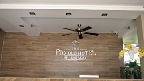 A reception area with a wooden wall, a ceiling fan, and a sign that reads "The Piccolo Hotel of Boracay".