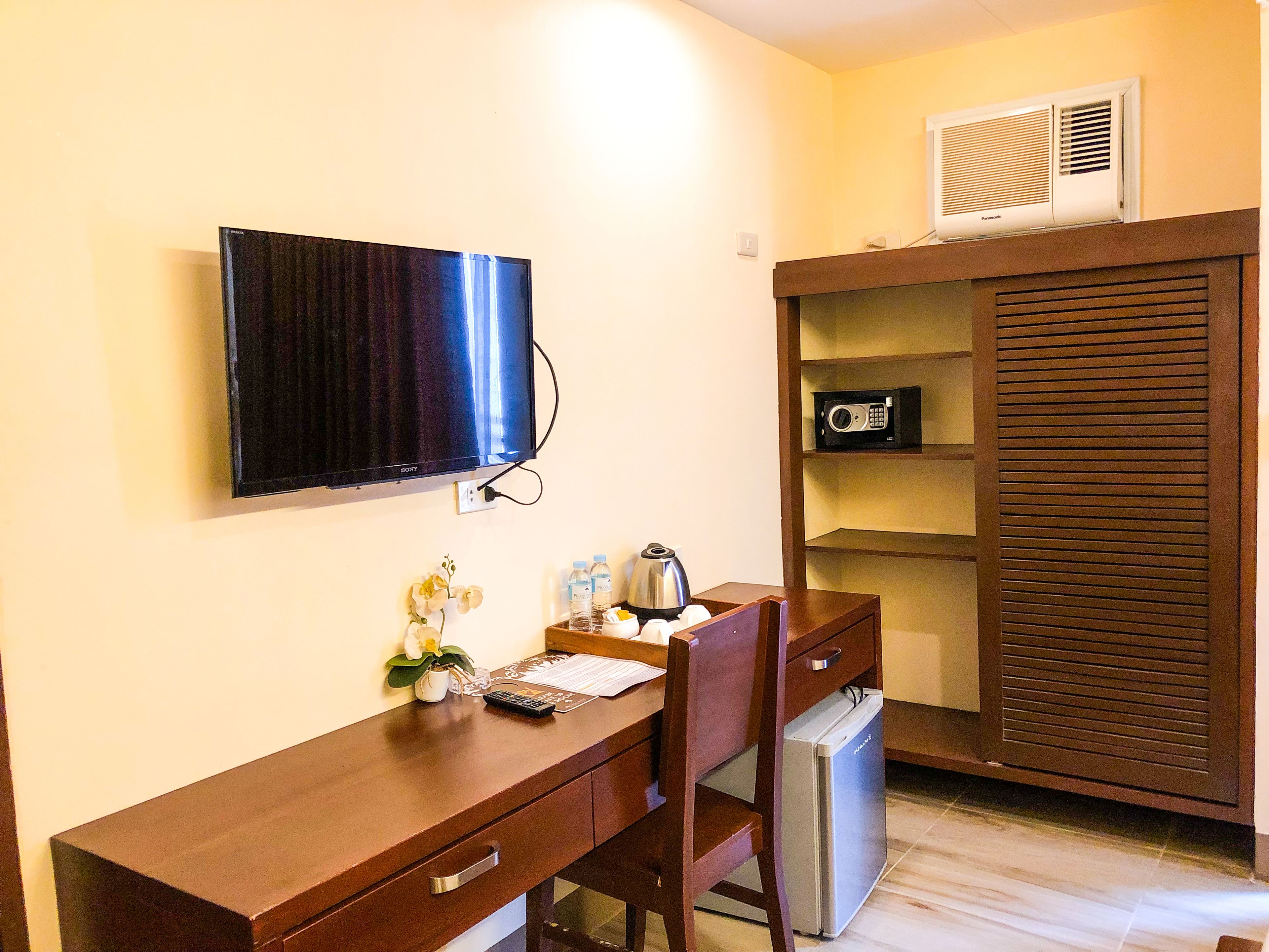 A compact hotel room with a wooden desk, a television mounted on the wall, a small refrigerator, and a built-in shelving unit with a microwave.
