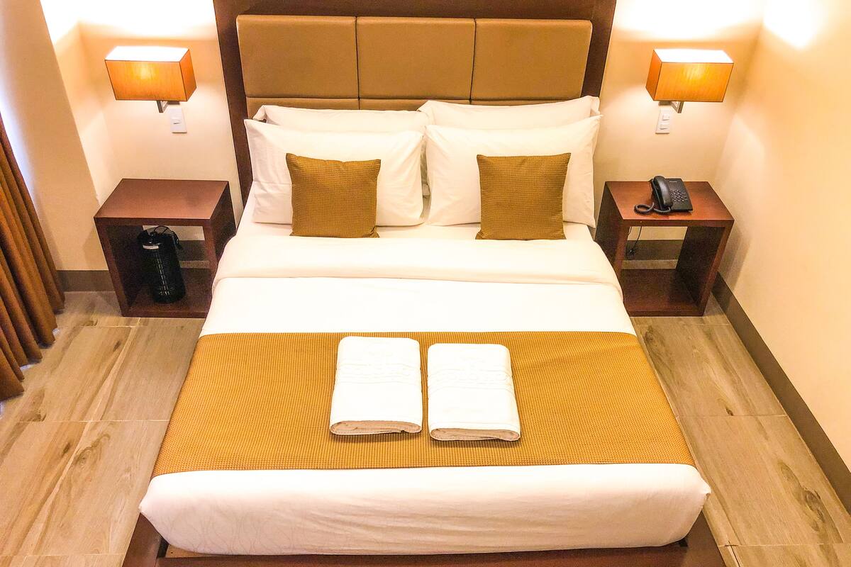 Una cama bien hecha con sábanas blancas y dos almohadas marrones, un cabecero de madera y dos lámparas de noche.
