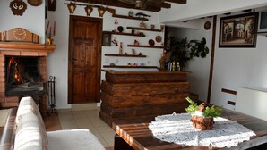 A cozy living room with a fireplace, wooden shelves, a dining table with a lace tablecloth, and a sofa. - Trenchova Guest House (Bansko)