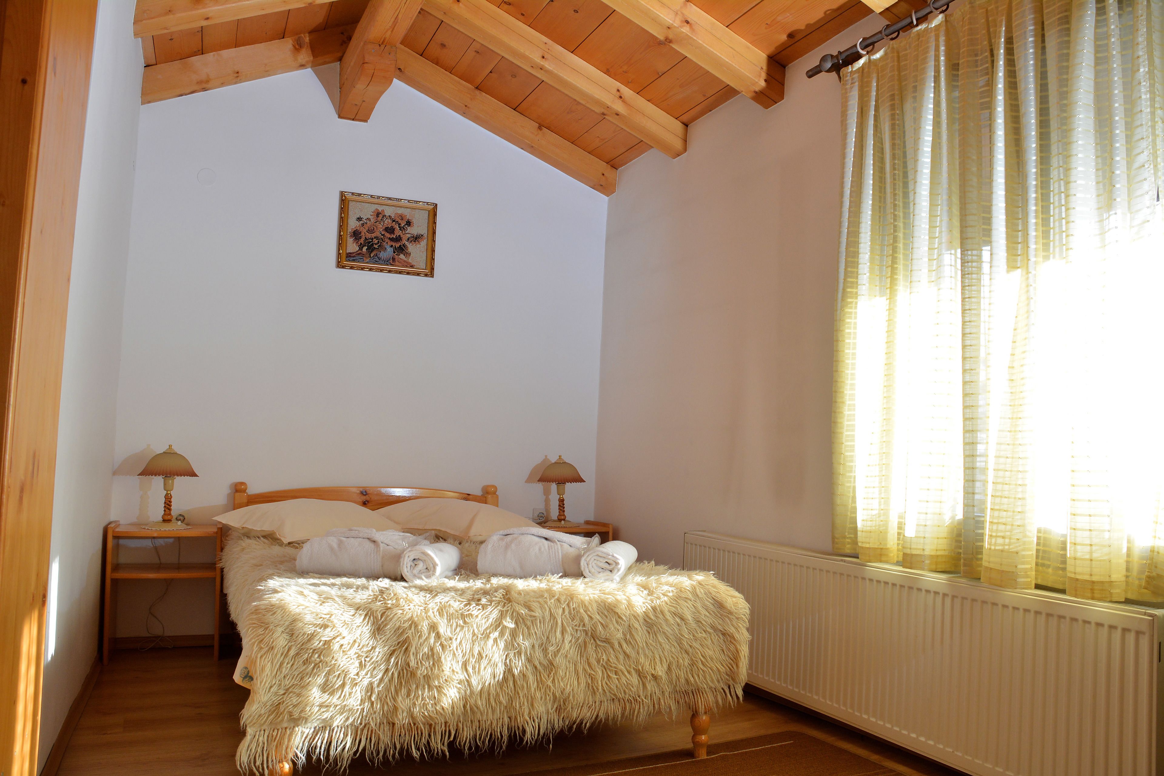 A bedroom with a wooden ceiling, a bed with a fluffy blanket, two bedside lamps, and a window with curtains.