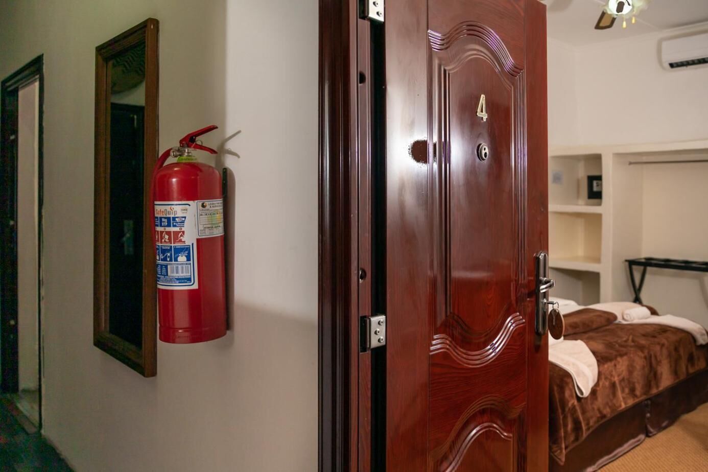 Een hotelkamer met een houten deur, een brandblusser aan de muur, een bed met een bruine deken en een nachtkastje.