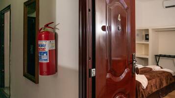 A hotel room with a wooden door, a fire extinguisher mounted on the wall, a bed with a brown blanket, and a bedside table.