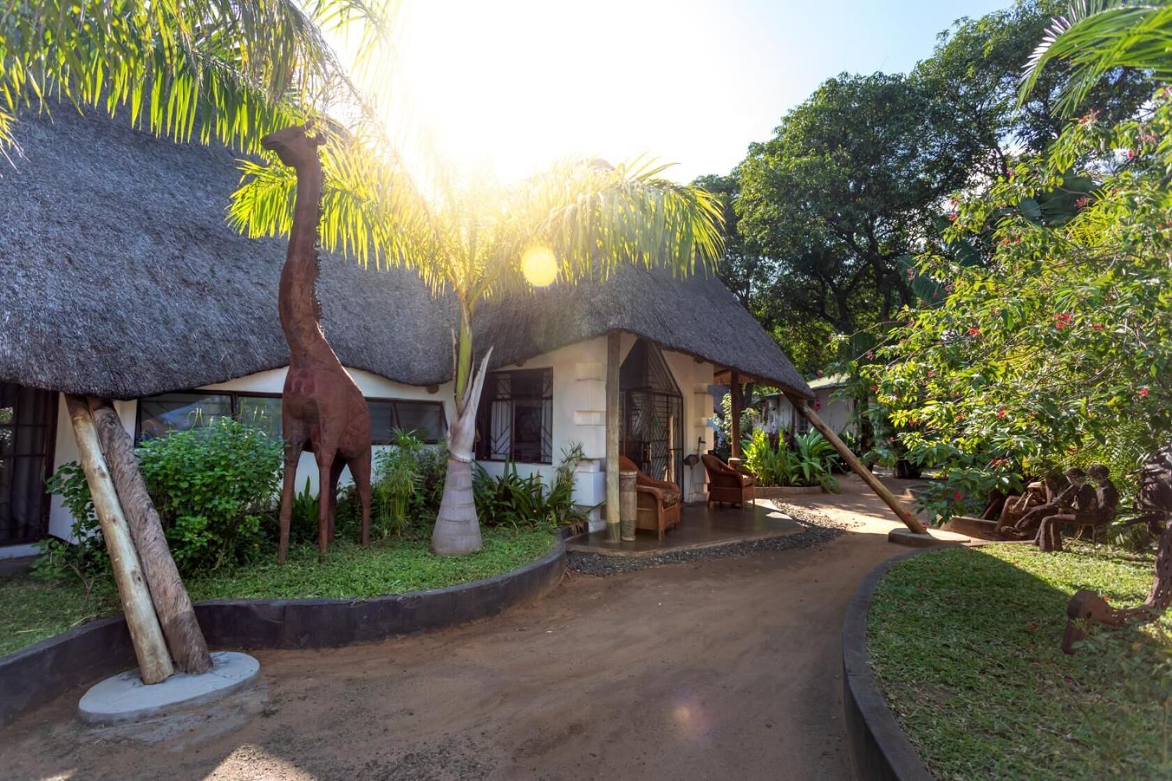 Un bâtiment à toit de chaume, avec un chemin de jardin qui y mène, entouré de plantes tropicales et orné d’une statue de girafe.