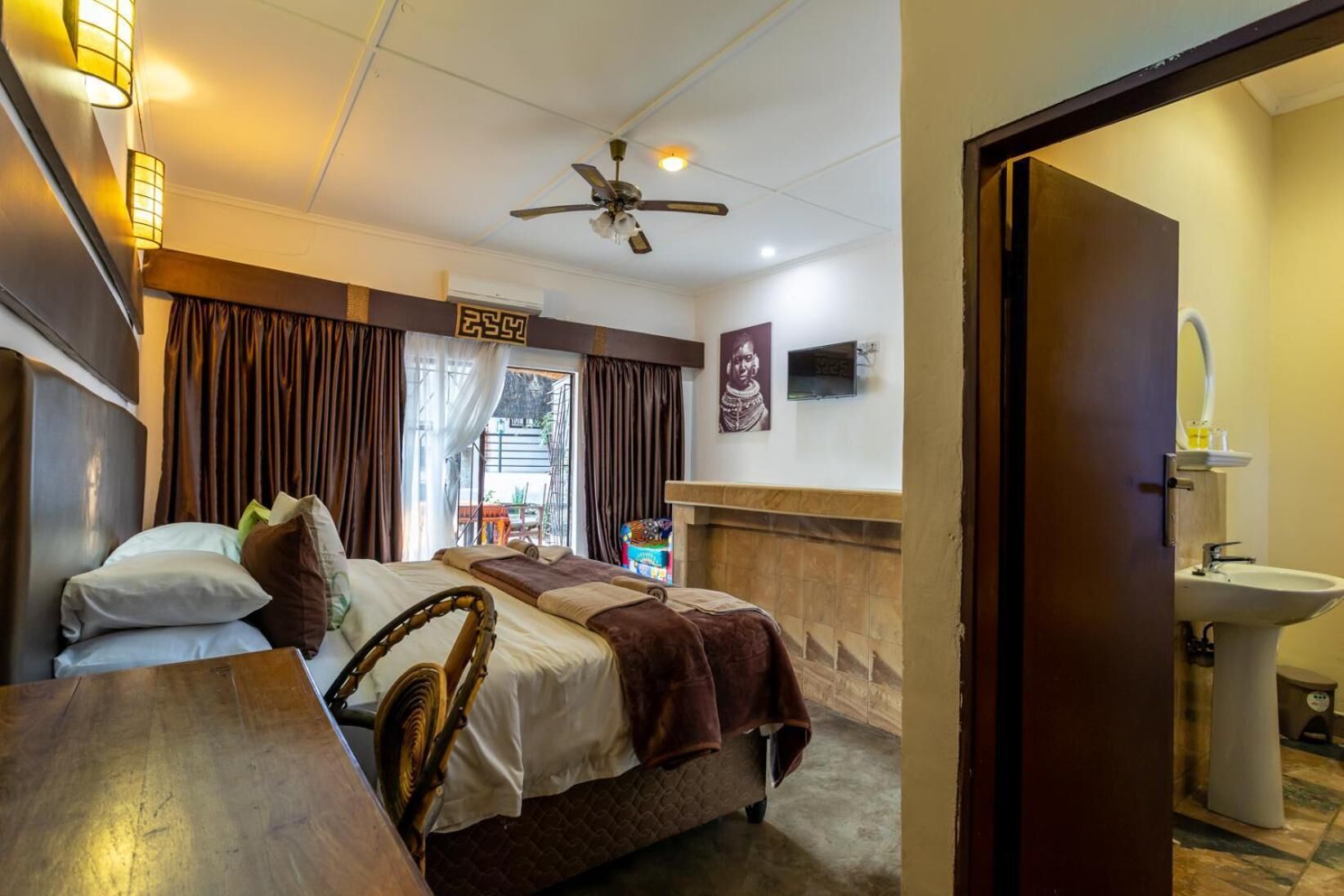 A hotel room with a bed, a wooden table, a ceiling fan, a window with curtains, a wall-mounted TV, and a bathroom with a sink and mirror.