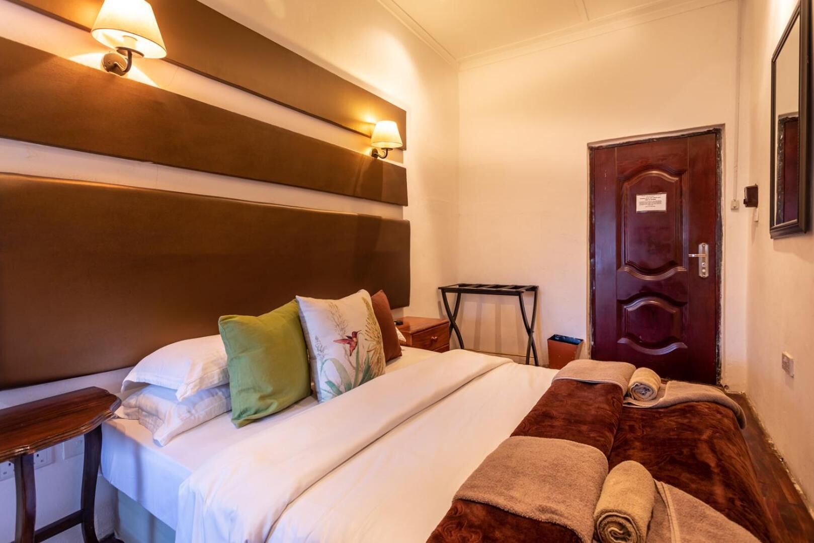 Habitación de hotel con una cama grande, cabecera de madera, dos lámparas fijadas a la pared, una puerta de madera y una pequeña mesa con una lámpara.