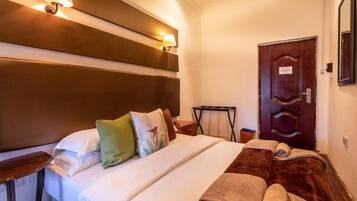Habitación de hotel con una cama grande, cabecera de madera, dos lámparas fijadas a la pared, una puerta de madera y una pequeña mesa con una lámpara.