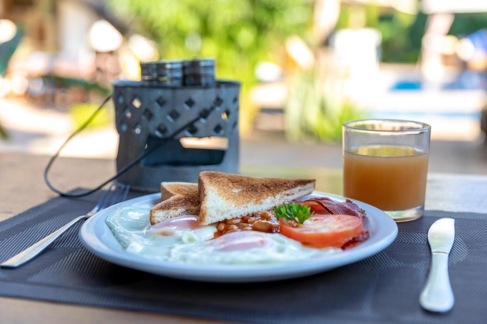Un plato de desayuno con tostadas, huevos, frijoles, tomate y tocino, acompañado de un vaso de jugo de naranja, sobre una mesa al aire libre.
