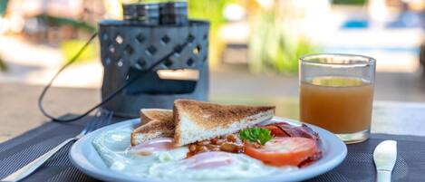 Un plato de desayuno con tostadas, huevos, frijoles, tomate y tocino, acompañado de un vaso de jugo de naranja, sobre una mesa al aire libre.
