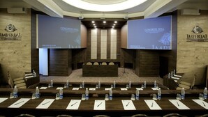 Una sala de conferències amb una taula llarga, cadires i un escenari. Hi ha dos pantalls grans a les parets que mostren el text "CONGRESS HALL".