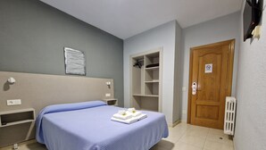 Habitación de hotel con cama, mesita de noche, estante, puerta y termostato.