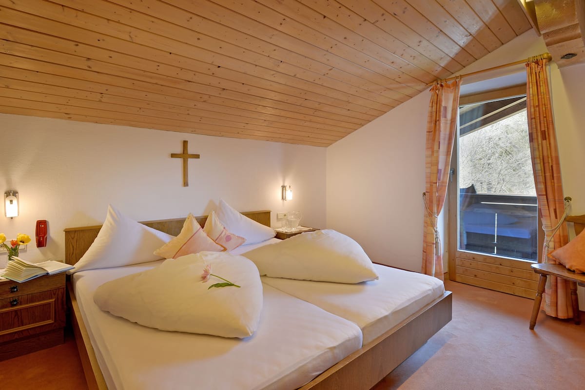 Una cama doble con ropa blanca, una cruz de madera en la pared y una ventana con cortinas.