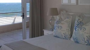 Un dormitorio con cama, mesita de noche, lámpara y vista al océano a través de un ventanal corredero.
