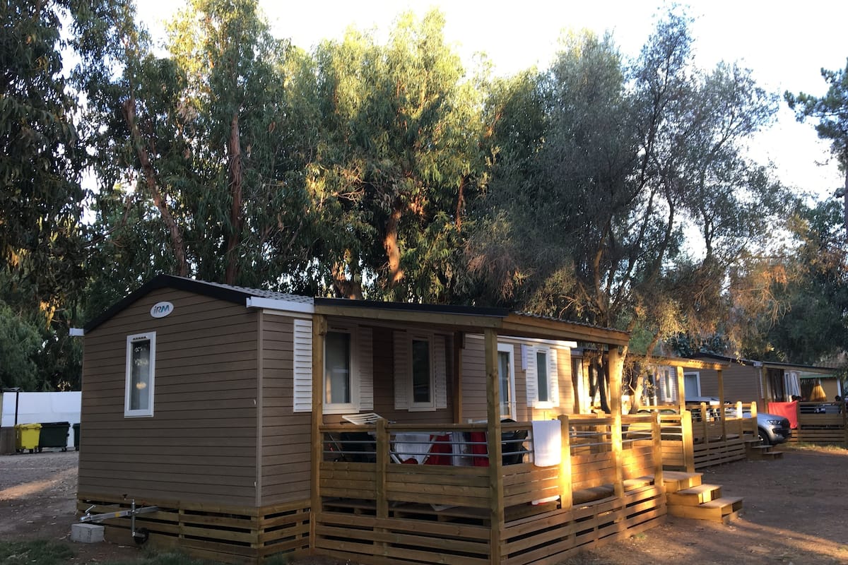Uma cabana de madeira pequena e térrea com varanda e área coberta, rodeada por árvores.