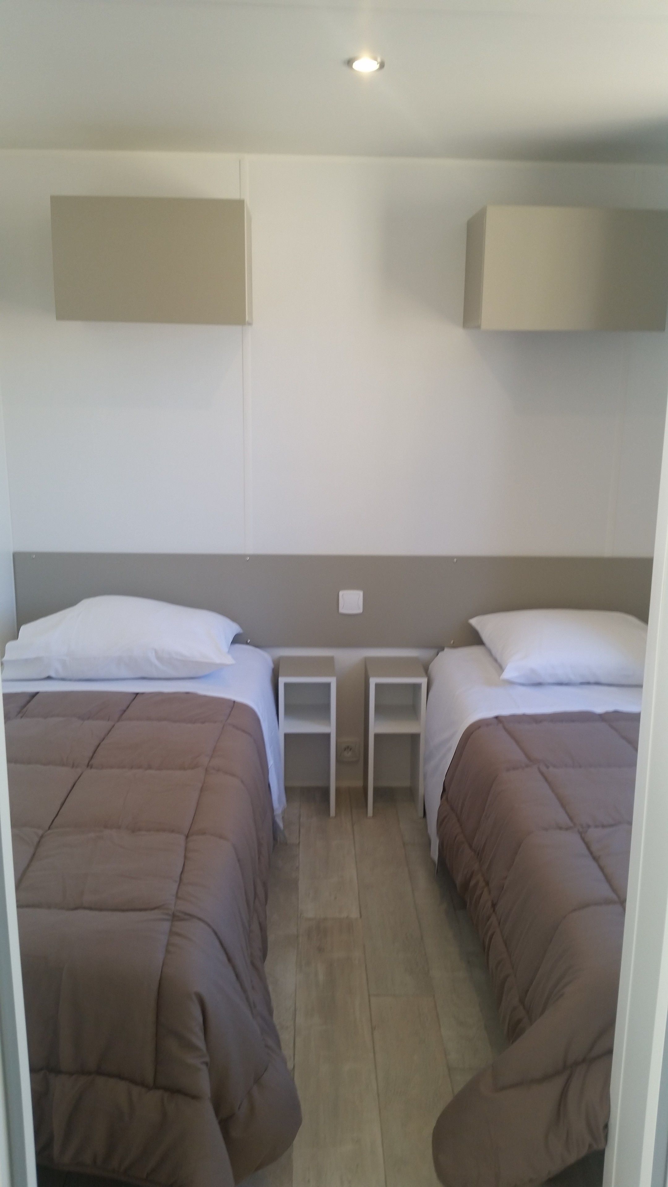 Habitación con dos camas, cada una con una almohada y un cabecero blanco. Hay dos mesitas de noche con lámparas. El piso y las paredes son de color claro, y se ve una lámpara en el techo.