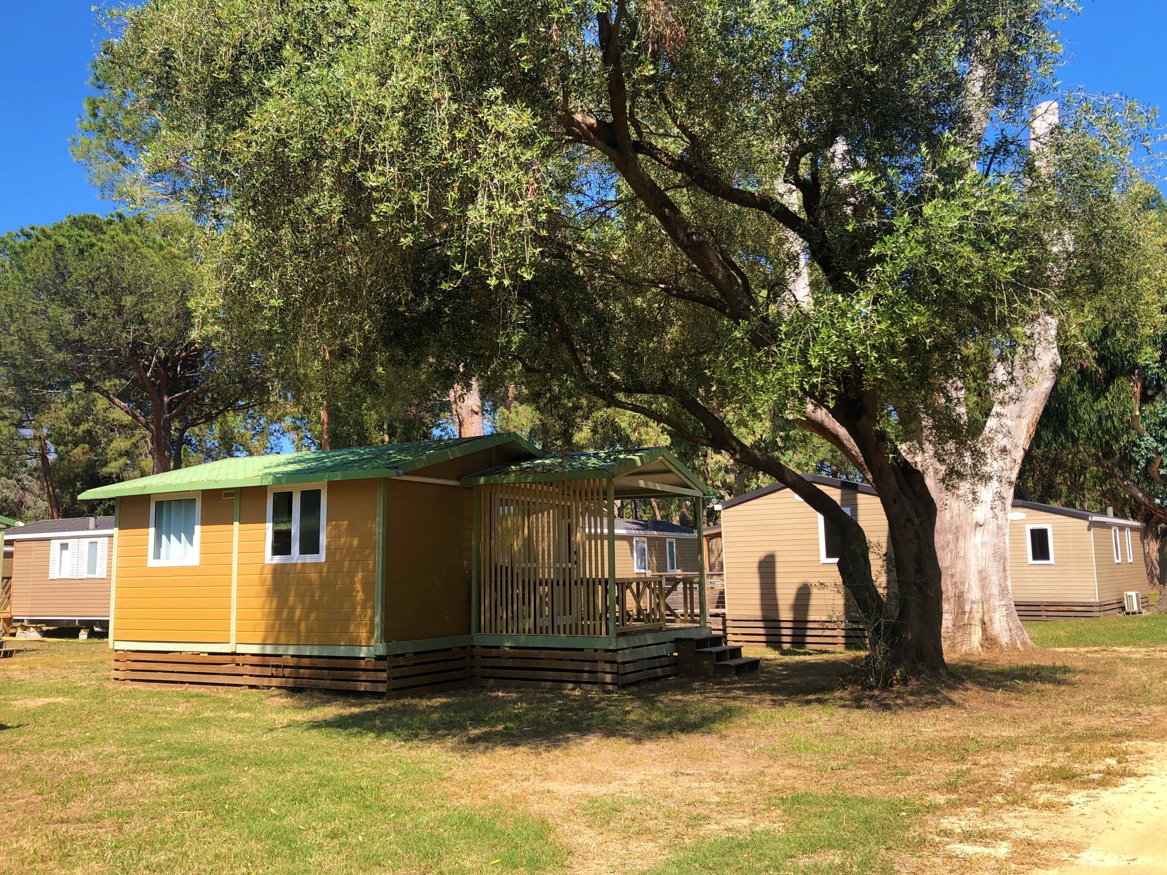 A small, single-story mobile home with a green roof and a wooden deck, surrounded by trees.
