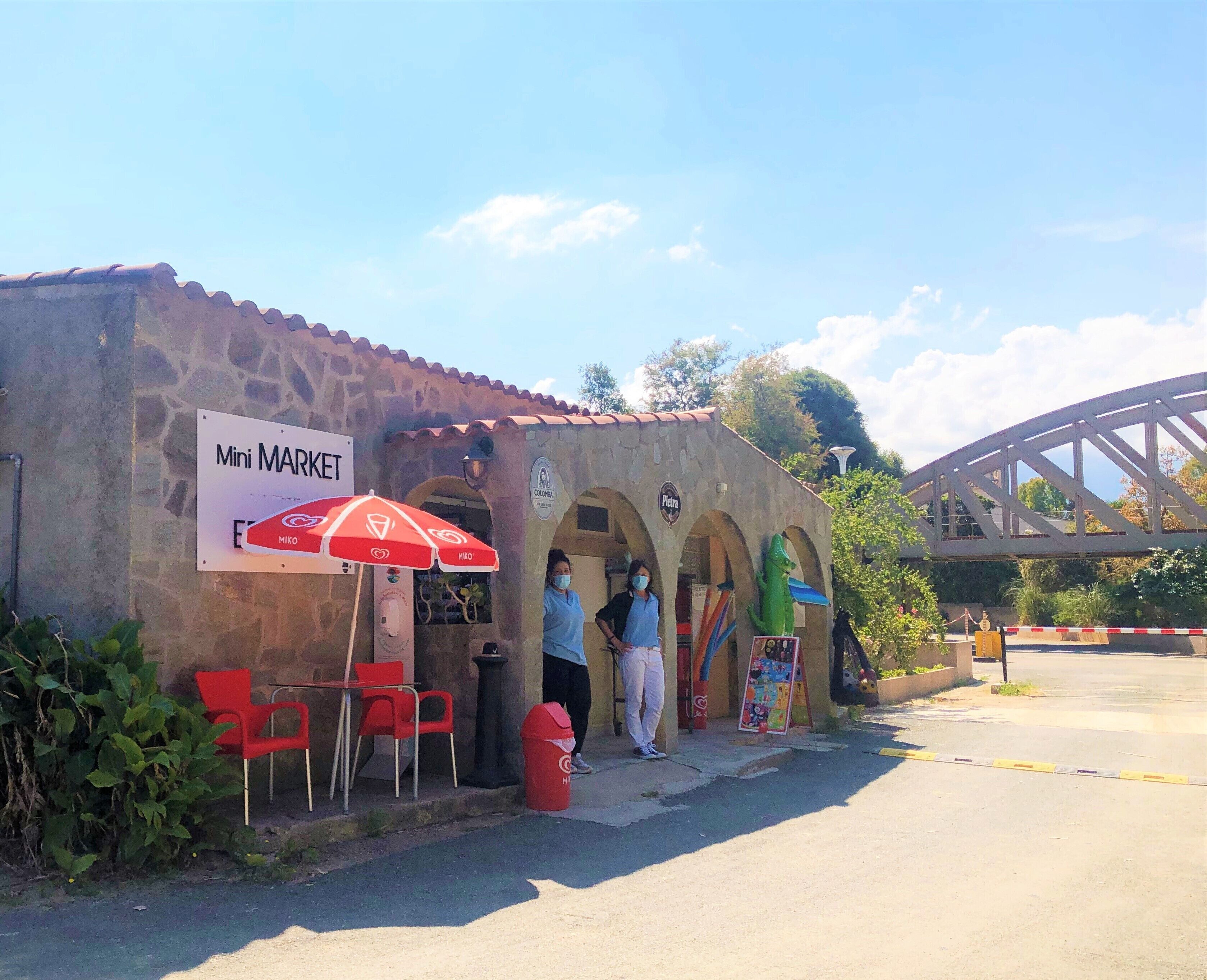 Un edificio de piedra con un letrero que dice "Mini MARKET" y un paraguas rojo. Hay sillas rojas afuera y un basurero.