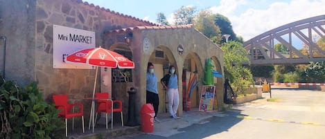 Un edificio de piedra con un letrero que dice "Mini MARKET" y un paraguas rojo. Hay sillas rojas afuera y un basurero.