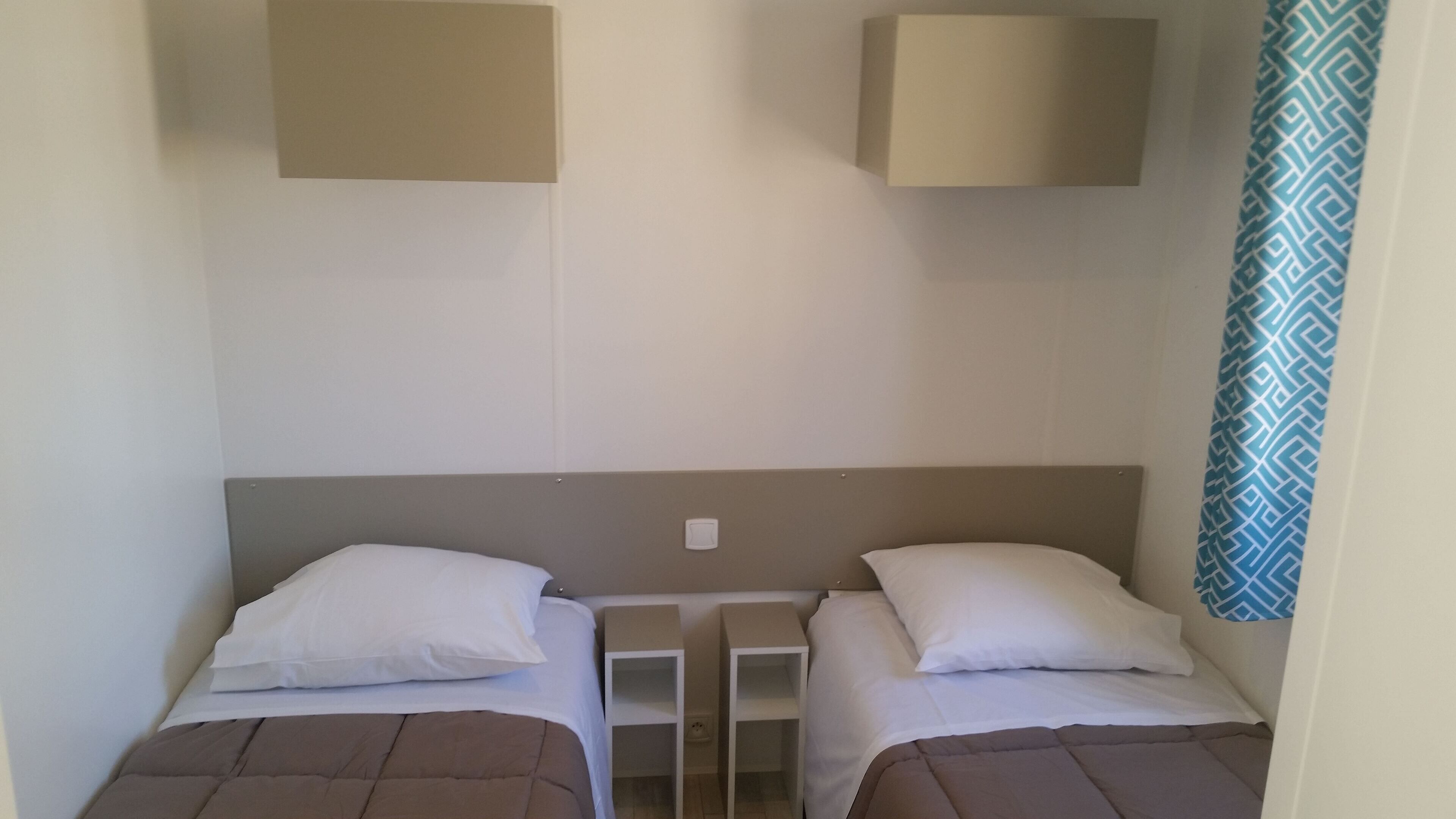A small, simple room with two single beds, each with a pillow and a brown blanket. There are two white nightstands between the beds, and a light switch on the wall above the beds.