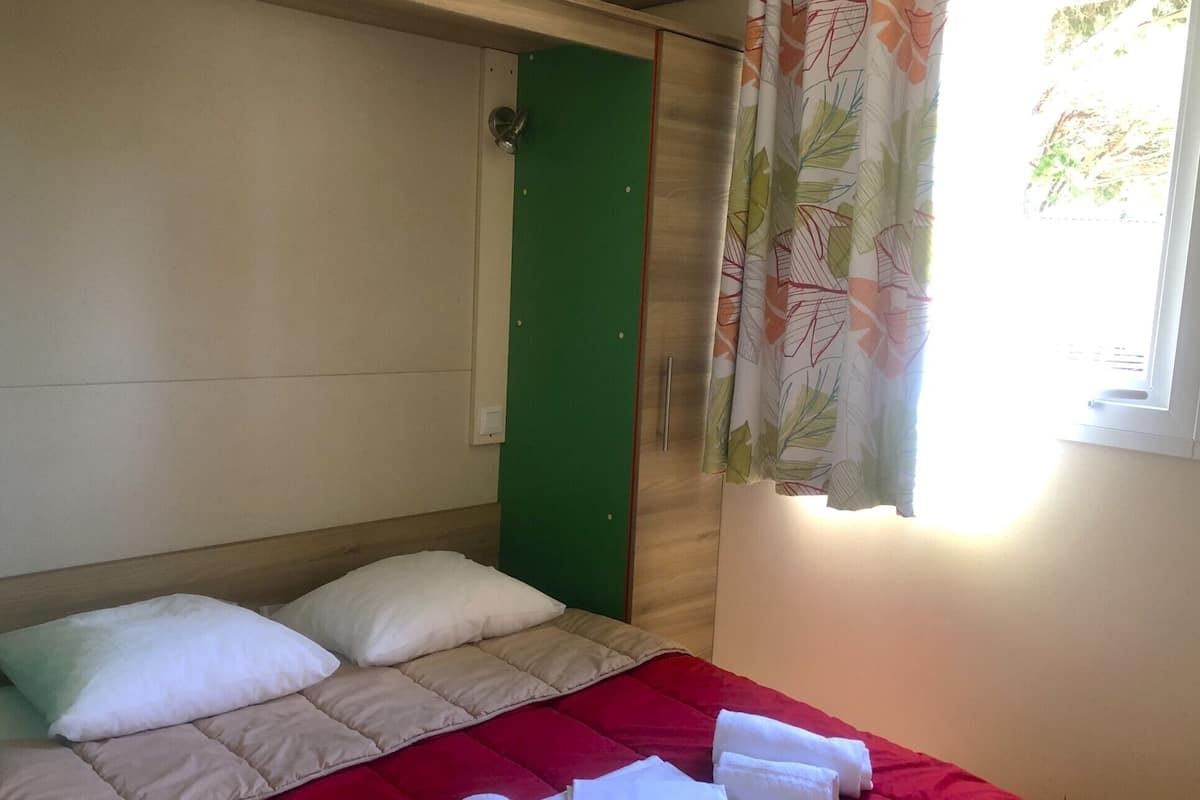 Uma cama de solteiro com colcha vermelha, duas almofadas e uma porta verde.