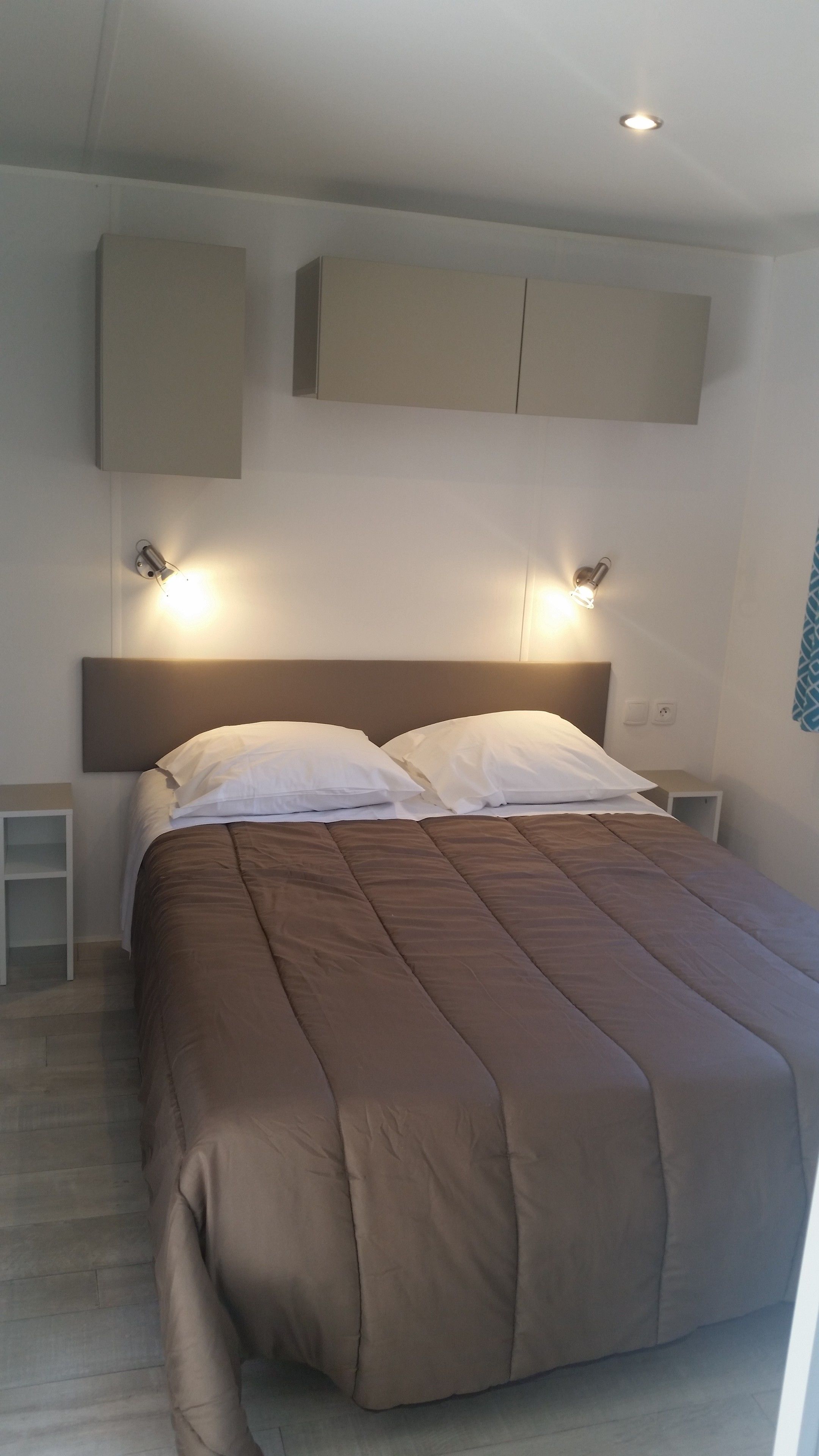 Ein ordentlich bezogenes Bett mit brauner Tagesdecke und weißen Kissen, zwei Nachttische mit Lampen und ein Wandregal über dem Bett.