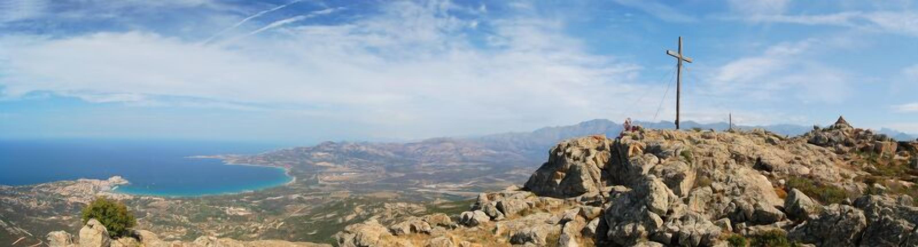 A rocky landscape with a cross on a hill, overlooking a coastal area with a clear sky.