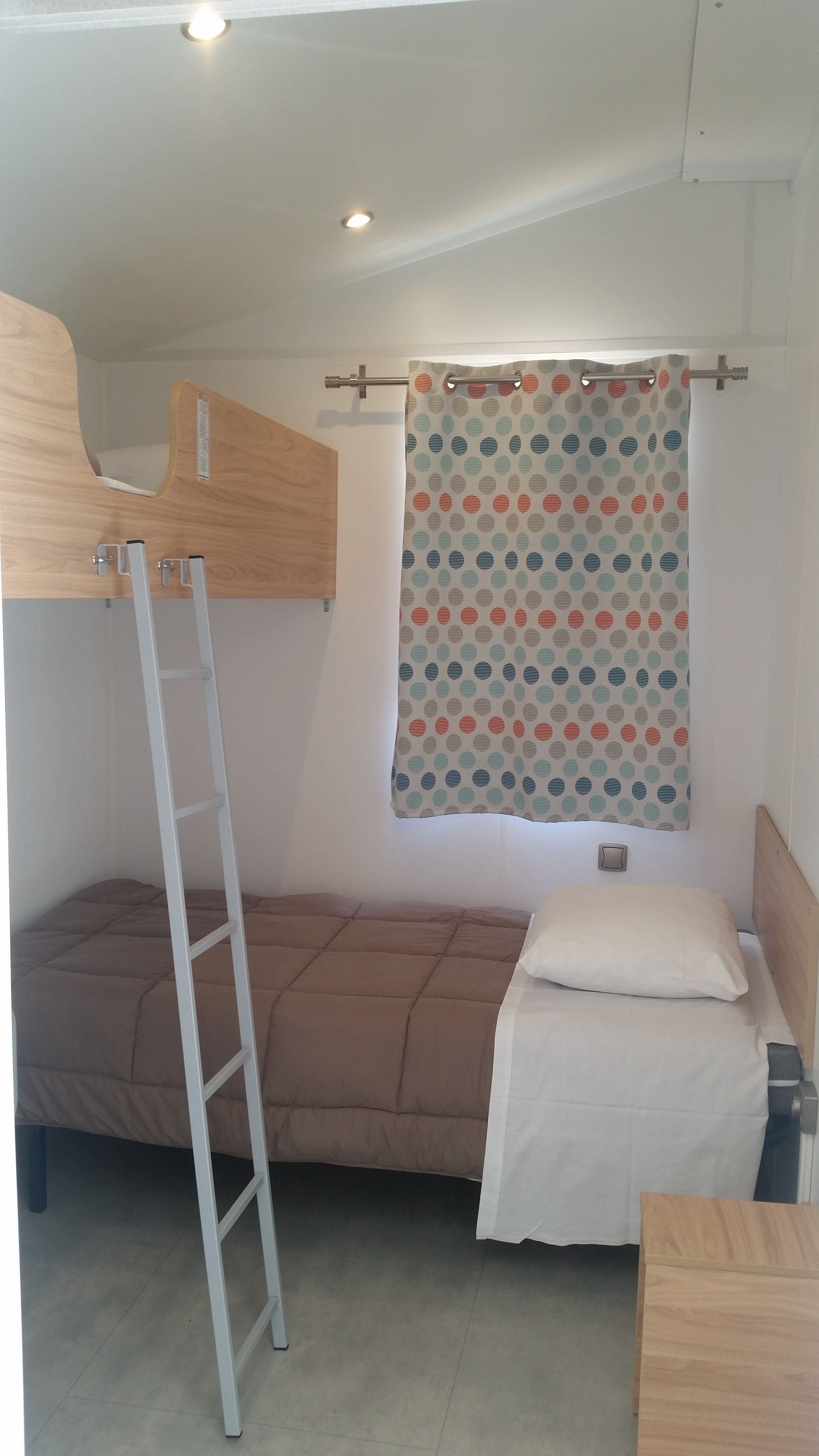 A compact room with a bunk bed, a small desk, and a window with a polka dot curtain.