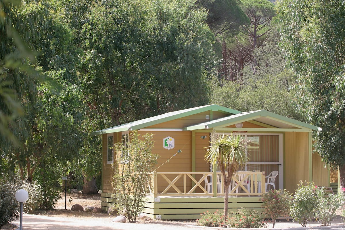 Um pequeno edifício térreo com telhado verde e varanda com cadeiras brancas.