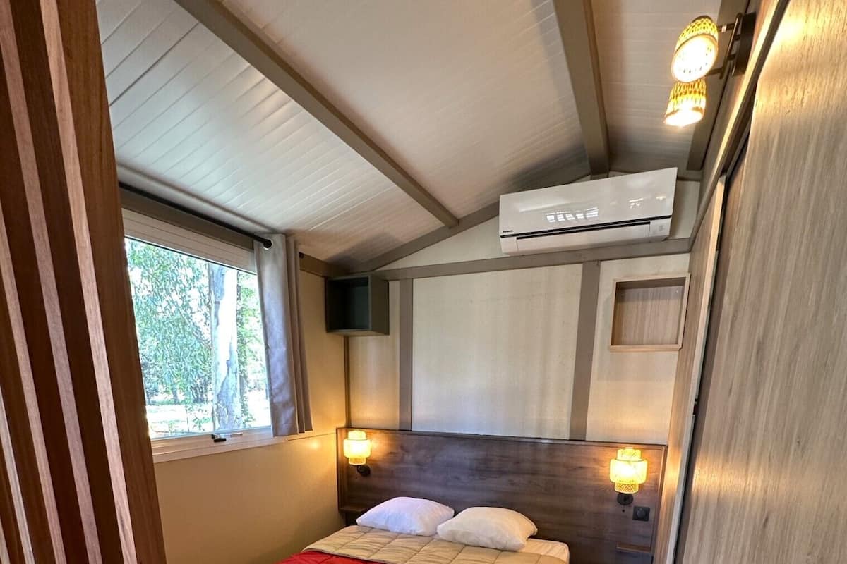 Quarto compacto com cama, luminárias de cabeceira, janela e ar condicionado.