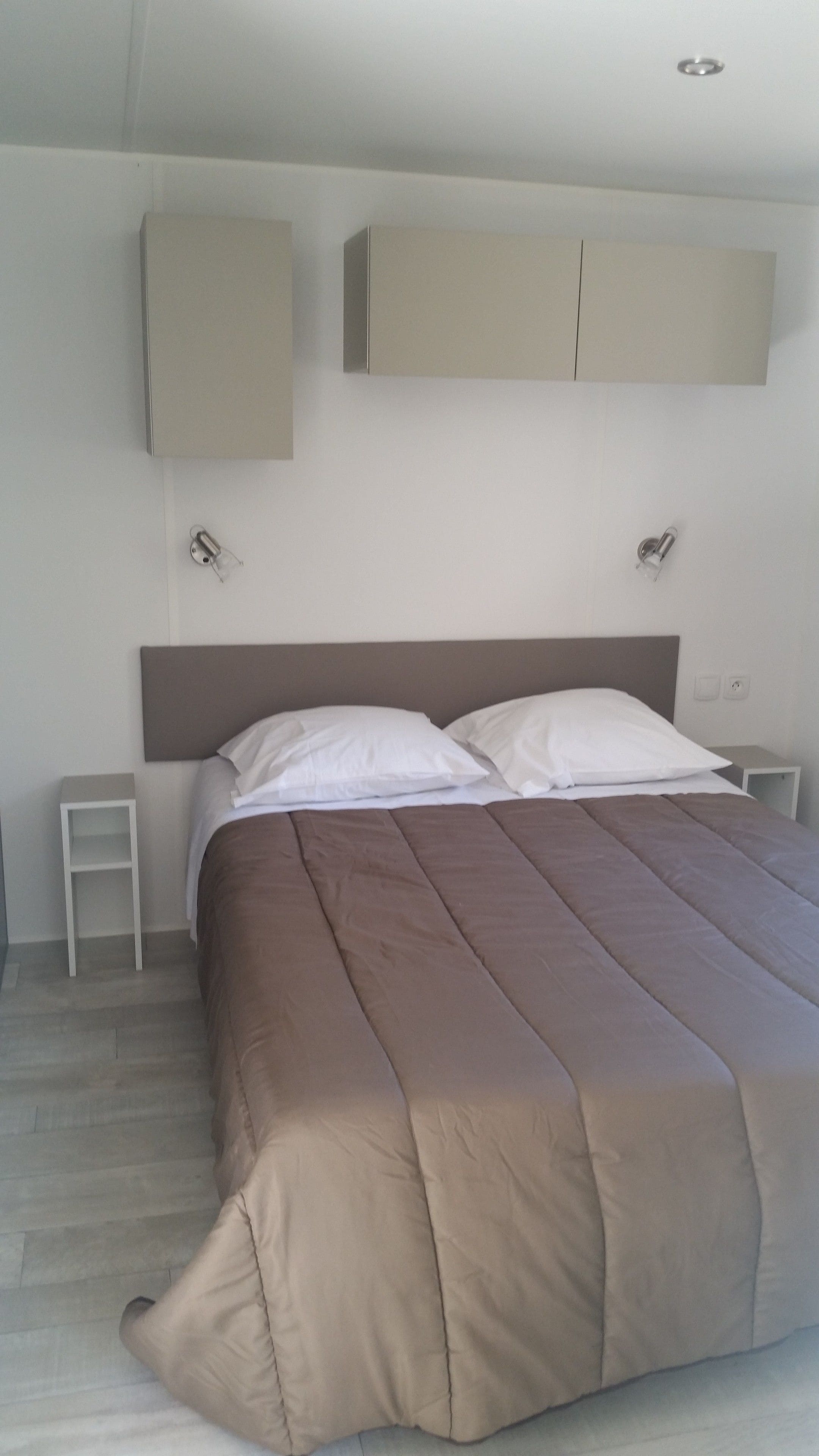Un lit bien fait, avec une couvre-lit marron et des oreillers blancs, une petite table de chevet et un meuble fixé au mur au-dessus du lit.