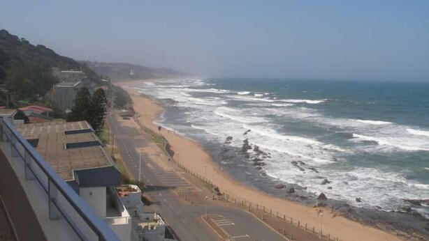 Eine Küstenstraße mit Strand und Meer rechts, Gebäuden links und wolkenlosem Himmel.