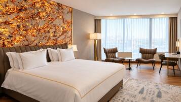 A hotel room with a large bed, two armchairs, a desk, and a view of the city through large windows.