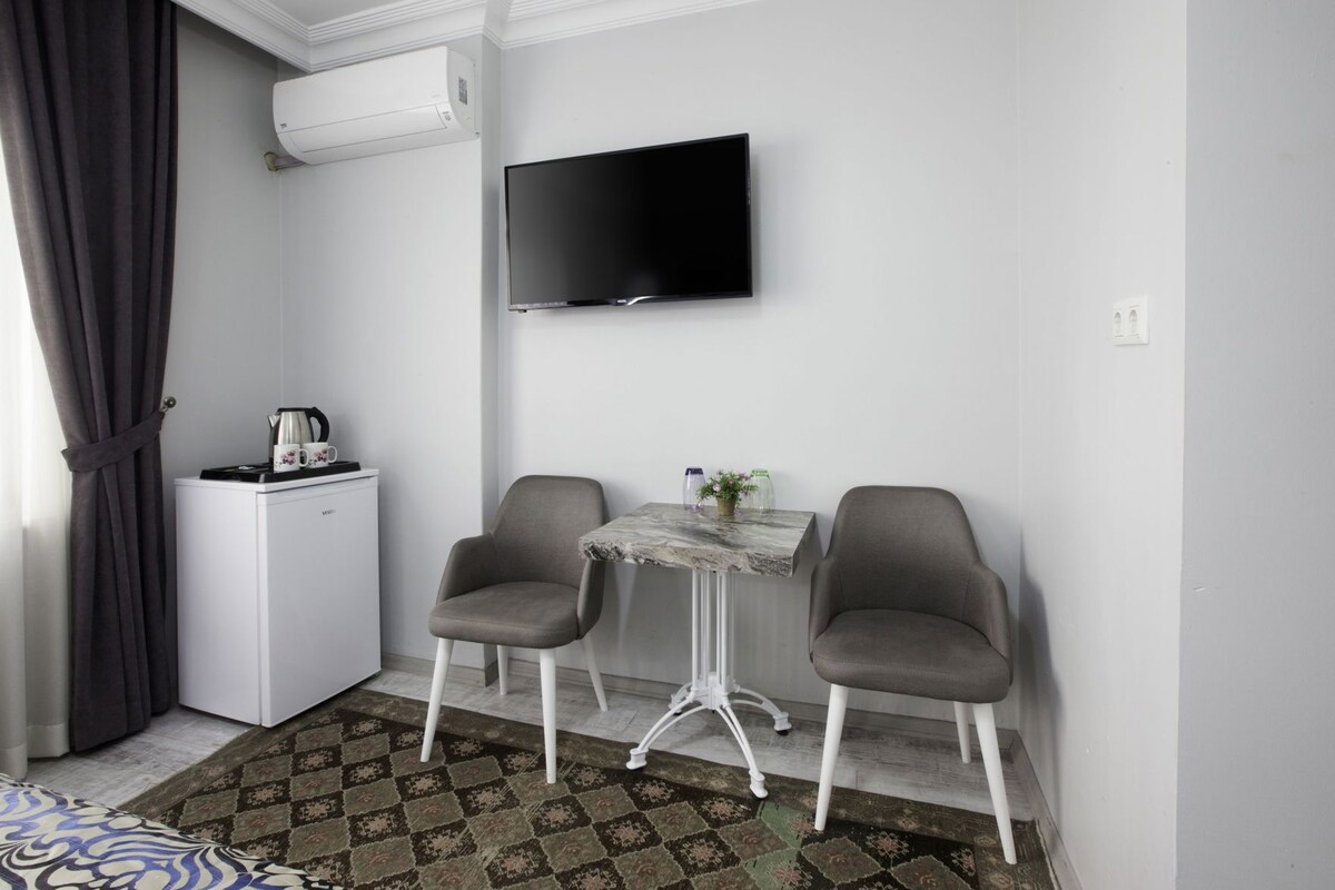 Um quarto pequeno com cama, escrivaninha, cadeira, televisão e ar condicionado.