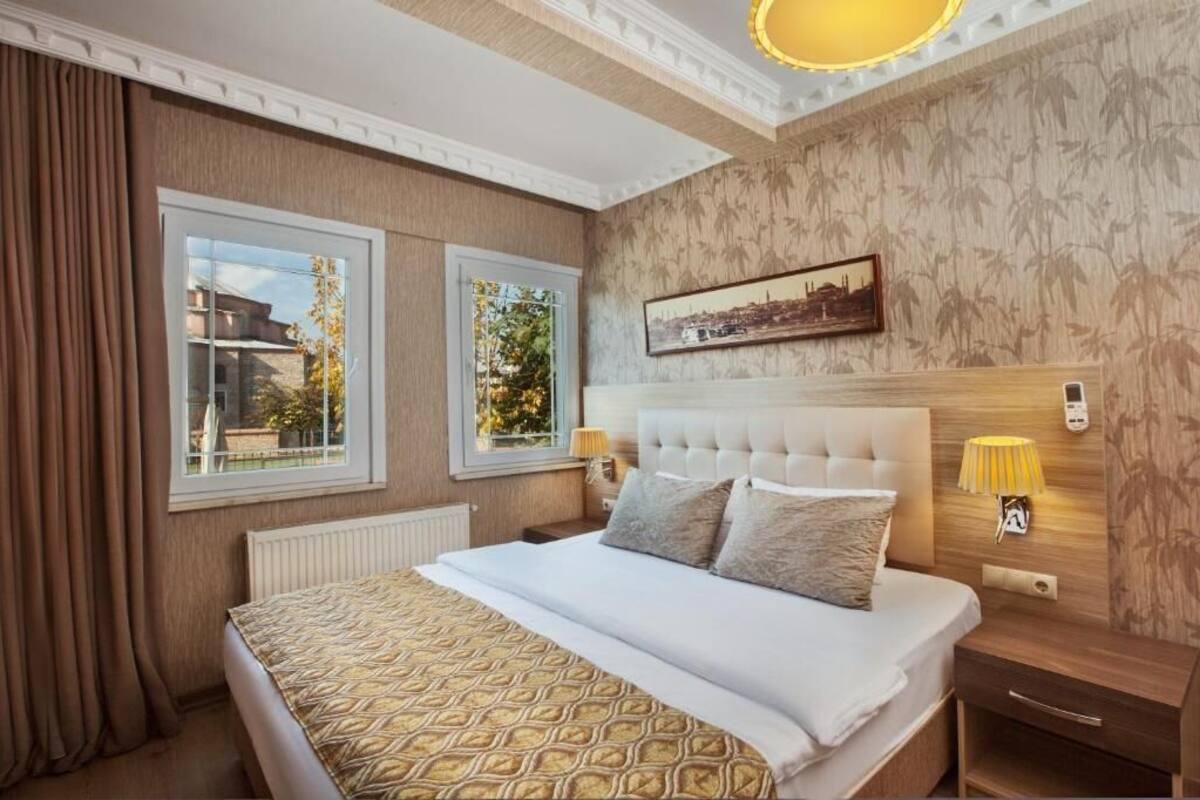 A hotel room with a large bed, two windows, a headboard with a cityscape painting, and a bedside lamp.