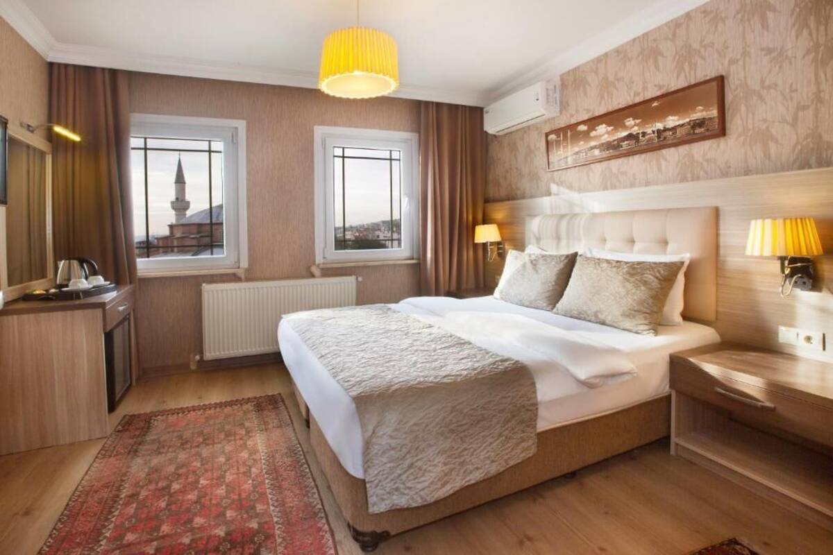 A hotel room with a large bed, two windows with views, a radiator, a bedside table with lamps, and a small desk with a kettle.