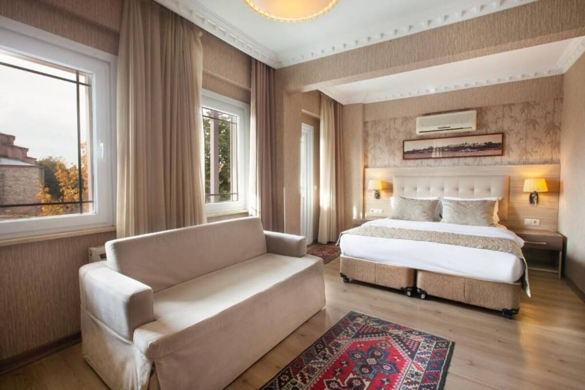 A hotel room with a large bed, a sofa, a patterned rug, and a view of the outside.