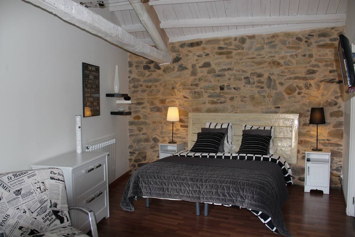 Un dormitorio con pared de piedra, una cama con colcha a rayas blanco y negro, dos lámparas de noche, un aparador blanco y una silla tapizada con estampado de periódico.