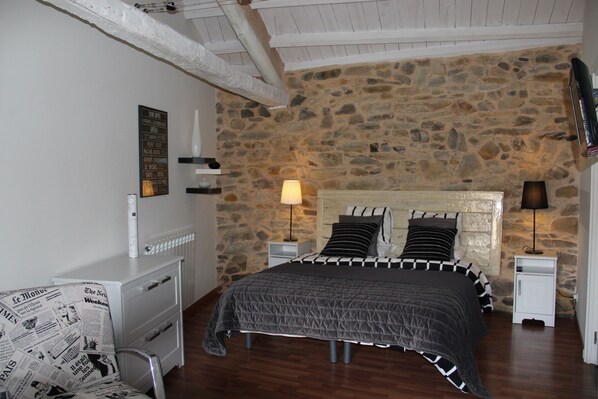 A bedroom with a stone wall, a bed with a black and white striped bedspread, two bedside lamps, a white dresser, and a chair with newspaper print upholstery.