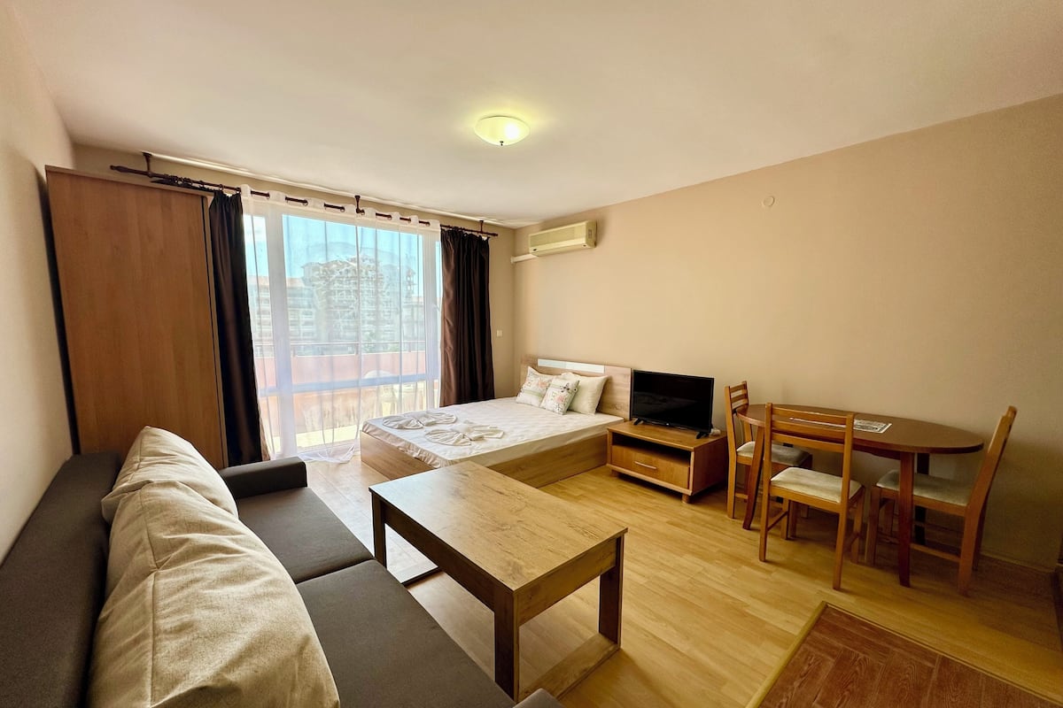 A modern hotel room with a sofa, a small dining table, a TV, and a balcony with curtains.